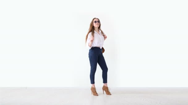 mode voor de moderne jeugd. stijlvolle meisje poseren tegen witte muur in jeans, wit t-shirt, met lederen rugzak en glazen. - Video
