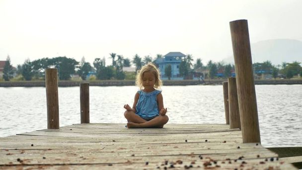 Child girl sits on the wood pier in lotus pose and meditating - 写真・画像