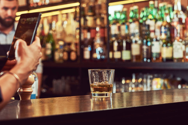 Cropped image of a customer of the bar takes a picture on the phone of a glass of alcohol on a bar counter. - Фото, изображение