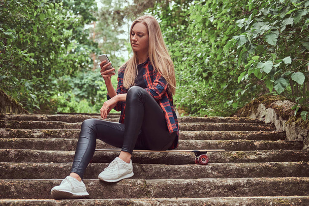 Young blonde hipster girl using a smartphone while sitting on steps in a park. - Fotó, kép
