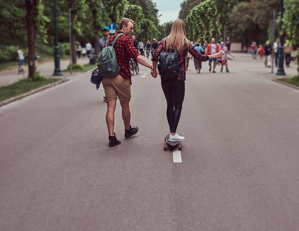 Young hipster couple. Guy teaches his girlfriend to skateboard on the street in a park. - Фото, зображення