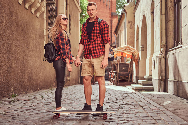 Young hipster couple, handsome skater and his girlfriend standing on old narrow streets of Europe. - Zdjęcie, obraz
