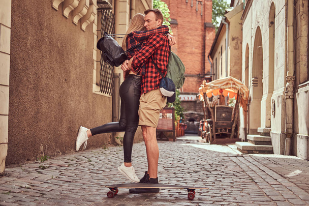Young hipster couple, handsome skater and his girlfriend cuddling while standing on the old narrow streets of Europe. - Photo, Image