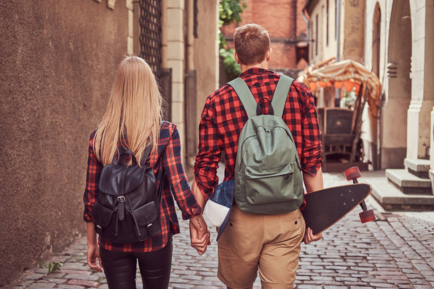 流行に敏感な若いカップル、ハンサムなスケーターとヨーロッパの古い狭い通りを歩いて、手をつないで、彼の恋人の背面図. - 写真・画像