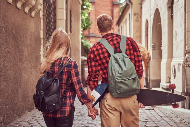 Back view of a young hipster couple, handsome skater and his girlfriend, holding hands, walking around a old narrow streets of Europe. - Photo, Image