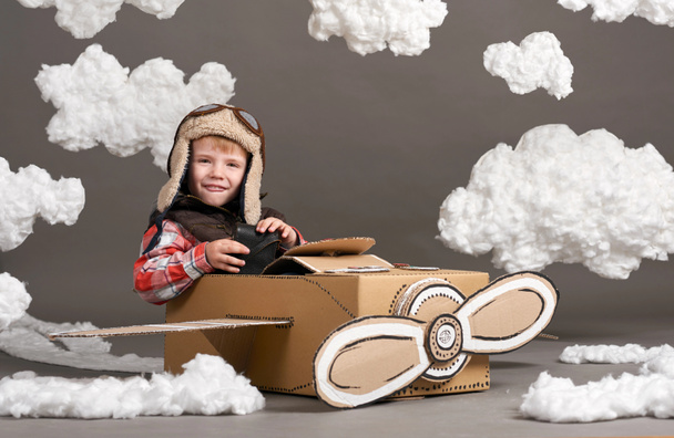 the boy plays in an airplane made of cardboard box and dreams of becoming a pilot, clouds of cotton wool on a gray background - Photo, Image