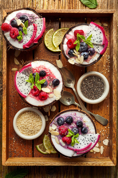Berry smoothie in a coconut bowl with fresh Pitaya fruit (Dragon fruit), blueberries and raspberries, mint, almond flakes, chia seeds and sesame seeds, top view. Healthy and delicious dessert or breakfast, vegetarian food - Photo, Image