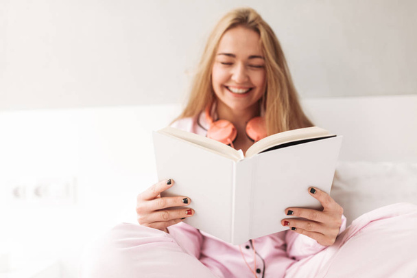 Close up photo of young smiling lady sitting on bed with book in hands and laughing at home isolated - Photo, Image