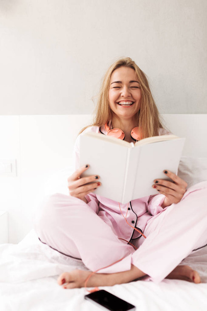 Молода красива леді сидить на ліжку з книгою в руках і сміється вдома ізольовано
 - Фото, зображення