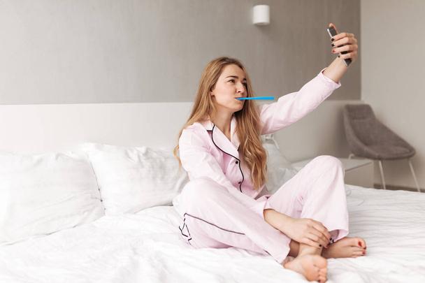 Retrato de jovem senhora bonito sentado na cama com escova de dentes e tirar fotos no celular em casa isolado
 - Foto, Imagem