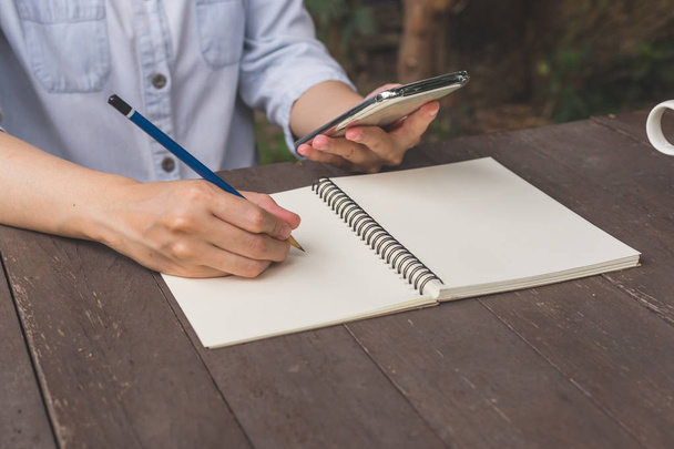 Kobieta ręka pisać notebook i trzymając telefon na stół z drewna. - Zdjęcie, obraz