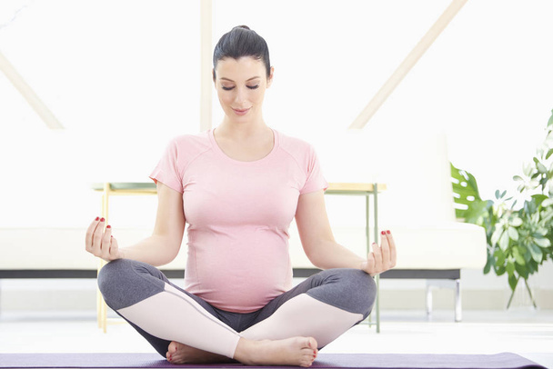 Full length shot of pregnant woman meditating in yoga pose at home.  - 写真・画像