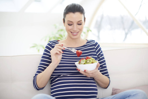 Portrait of beautiful pregnant woman relaxing at home and eating a bowl of fruit salad. - Foto, Imagem