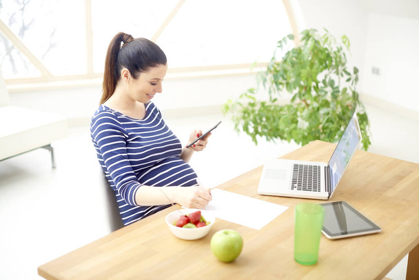Smiling pregnant woman text messaging and writing something while sitting behind the computer and working from home.  - 写真・画像