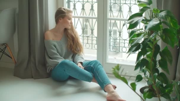 Young pensive woman sitting by a window. - Filmati, video