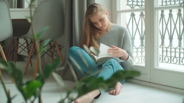 Young woman at home sitting near a window reading book and relaxing. - Záběry, video