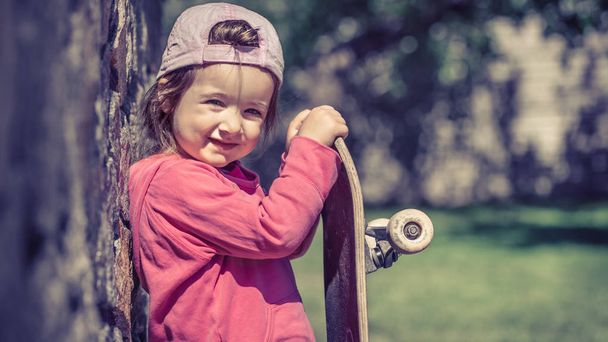 ein modisches kleines Mädchen hält ein Skateboard in der Hand und spielt - Foto, Bild