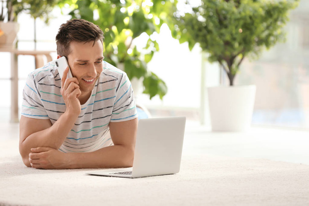 Handsome young man with mobile phone and laptop on cozy carpet at home - Foto, Imagem