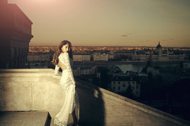 Woman in white wedding dress on evening city view, fashion. Sensual woman with long hair on balcony, beauty. Bride with makeup. Girl with glamour look in wedding - Φωτογραφία, εικόνα
