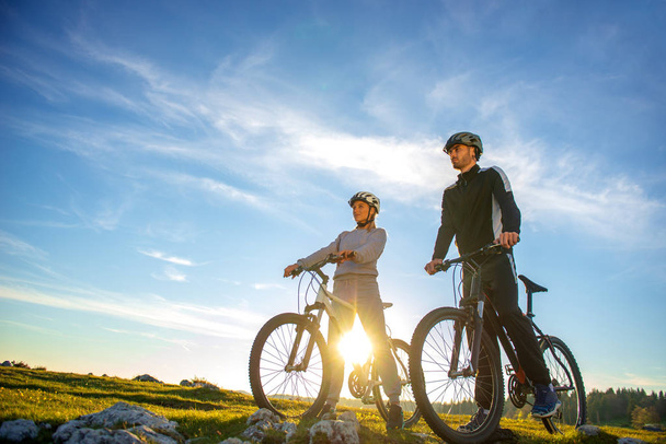 Ciclista casal com bicicletas de montanha em pé na colina sob o céu à noite e desfrutar de sol brilhante ao pôr do sol
. - Foto, Imagem