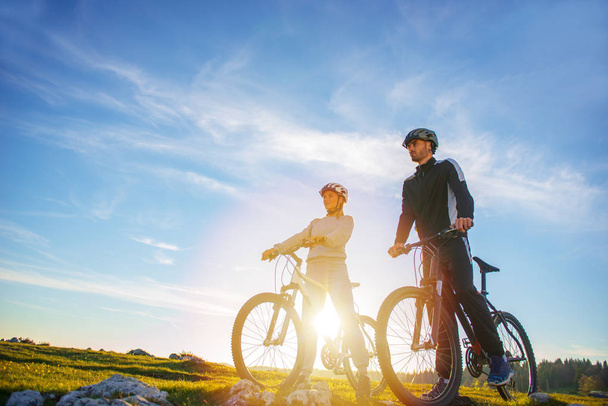 Radfahrerpaar mit Mountainbikes steht auf dem Hügel unter dem Abendhimmel und genießt die strahlende Sonne beim Sonnenuntergang. - Foto, Bild
