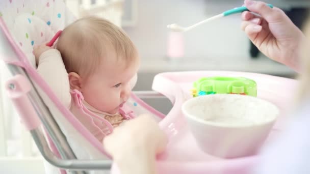 Baby with toy sitting in chair and eating puree. Mother feeding baby with spoon - Záběry, video