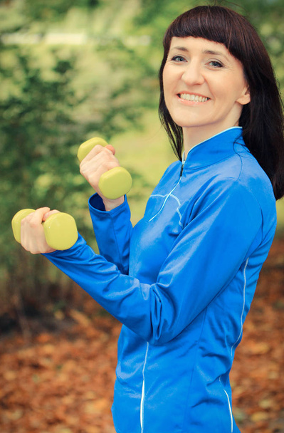 Smiling woman in sporty clothes exercising with dumbbells in park - Valokuva, kuva