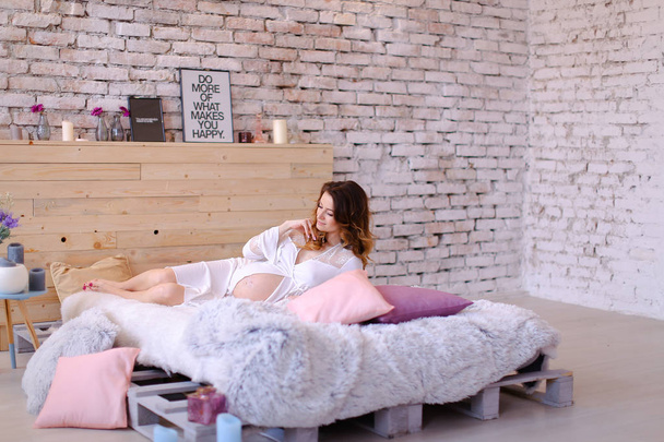 Pregnant young woman lying on bed in bedroom, wearing white dress. - Photo, Image