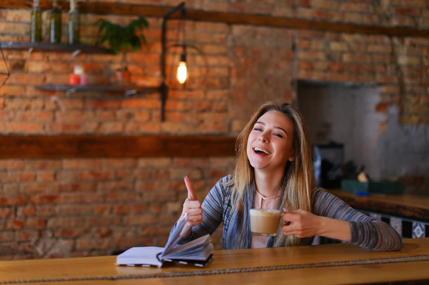 Piękna dziewczyna siedzi w pobliżu tabeli z notebooka, trzymając kubek kawy i śmiejąc się. - Zdjęcie, obraz