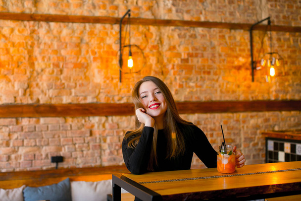 Tyytyväinen tyttö istuu lähellä puinen pöytä kuppi limonadia kahvilassa ja rentouttava
. - Valokuva, kuva