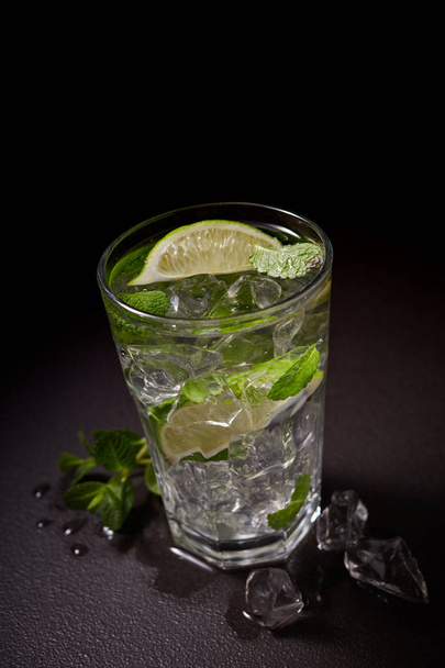Glass of iced Mojito cocktail and ingredients,  lime, ice and mint leaves on black background, cold summer drink  - 写真・画像