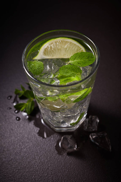 Glass of iced Mojito cocktail and ingredients,  lime, ice and mint leaves on black background, cold summer drink  - Photo, Image
