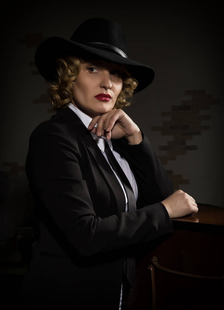 Elegant woman in business suit with a hat poses on a dark background, stylized retro portrait - Fotó, kép