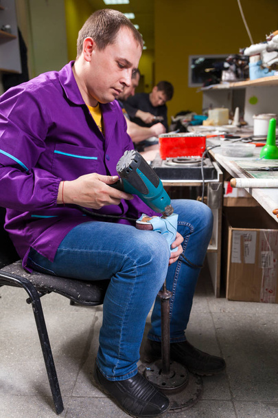 Un joven trabajador en jeans hace una suela de zapatos para niños de cuero genuino con un secador de pelo especial en el taller de costura
 - Foto, imagen