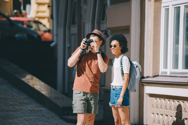 homem viajante tirar foto na câmera e Africano americano namorada de pé perto
  - Foto, Imagem