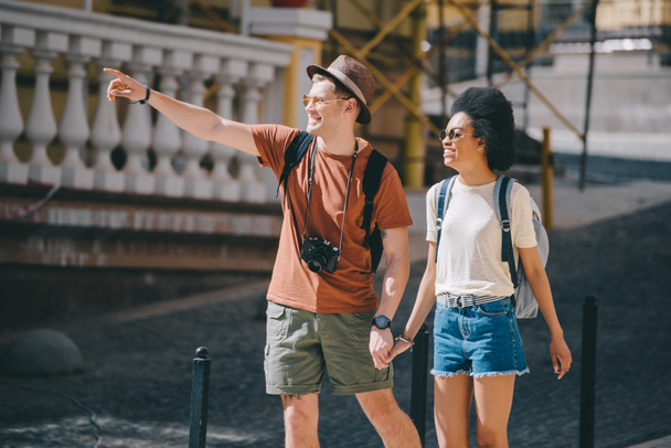 male traveler with camera pointing by finger to african american girlfriend walking near  - Photo, Image