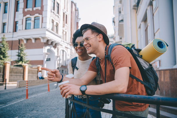 mladí usmívající se muž turistovi šálek ukazující prstem na africké americké přítelkyni  - Fotografie, Obrázek