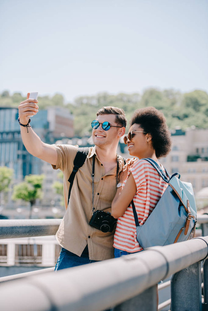 happy multicultural couple of tourists in sunglasses taking selfie on smartphone  - Zdjęcie, obraz