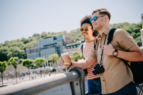 sonriente interracial pareja de turistas en gafas de sol con café y cámara mirando a la vista desde el puente
  - Foto, Imagen