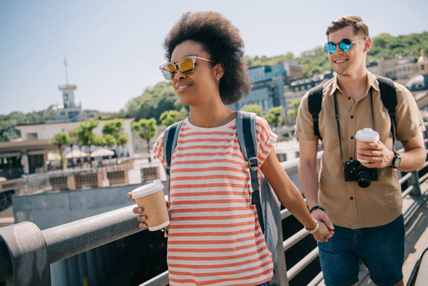 interracial couple of tourists with paper cups of coffee and camera holding hands and walking on bridge - Photo, Image