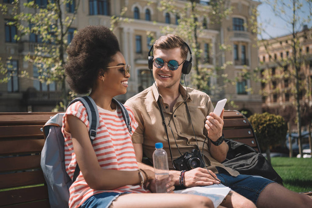 stylish couple with camera, headphones and smartphone sitting on bench  - Foto, imagen