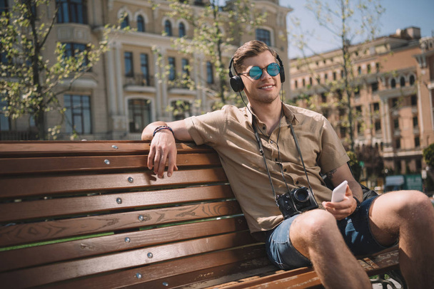 young man in sunglasses with camera, headphones and smartphone sitting on bench  - Fotografie, Obrázek