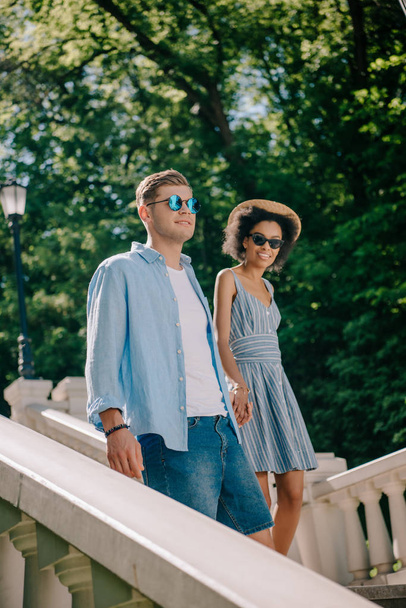 smiling interracial couple in sunglasses going downstairs in park  - Φωτογραφία, εικόνα