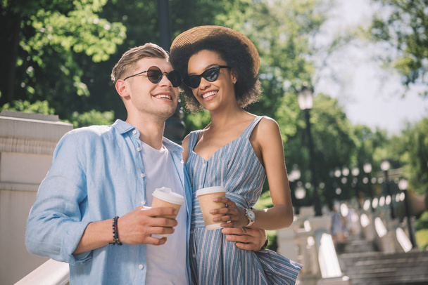 happy multiethnic couple in sunglasses with coffee cups standing on stairs in park  - Foto, imagen