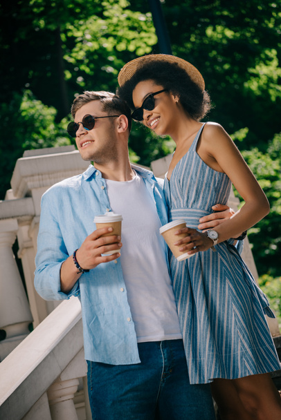 casal elegante interracial em óculos de sol com copos de papel de café nas escadas
  - Foto, Imagem