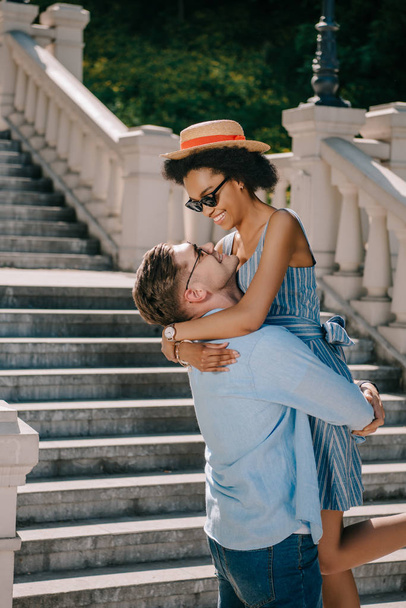 smiling young man holding african american girlfriend near stairs in park  - Photo, Image