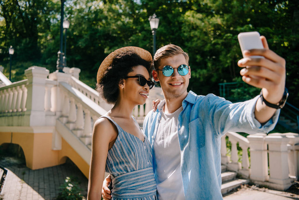 sonriente pareja interracial en gafas de sol tomando selfie en smartphone en parque
  - Foto, Imagen