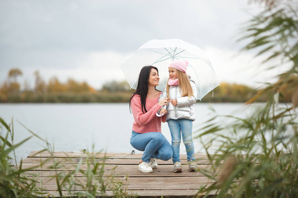 Jovem mãe com sua filhinha no tempo de outono perto do lago
 - Foto, Imagem