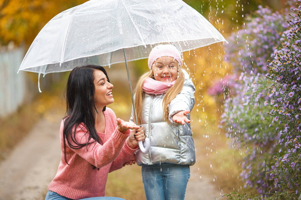 Adorable child with her young mother having fun in autumn background under the umbrella. Cheerful family in the fall time - Foto, afbeelding