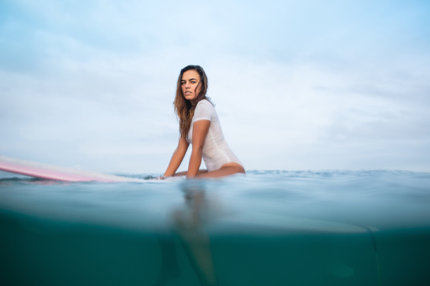 young woman in wet white swimsuit sitting sitting on surfboard in ocean and looking at camera - Photo, Image